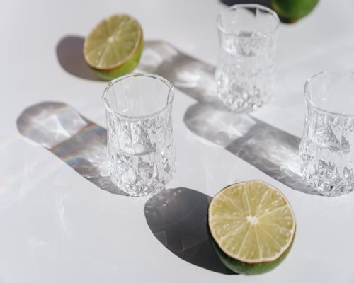 A sleek glass water container filled with fresh crystal clear water sitting next to cut lime slices on a table under natural sunlight shadow